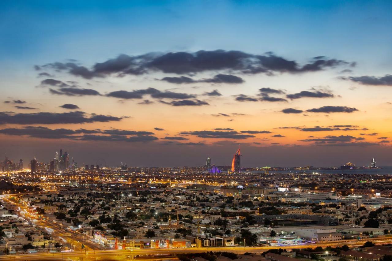 두바이 밀레니엄 아트리아 비즈니스 베이 아파트 호텔 외부 사진 The photo shows a stunning cityscape at dusk, with a panoramic view of a city skyline. The sky is transitioning from blue to orange, indicating sunset, with scattered clouds creating a colorful backdrop. In the foreground, you can see a sprawling urb