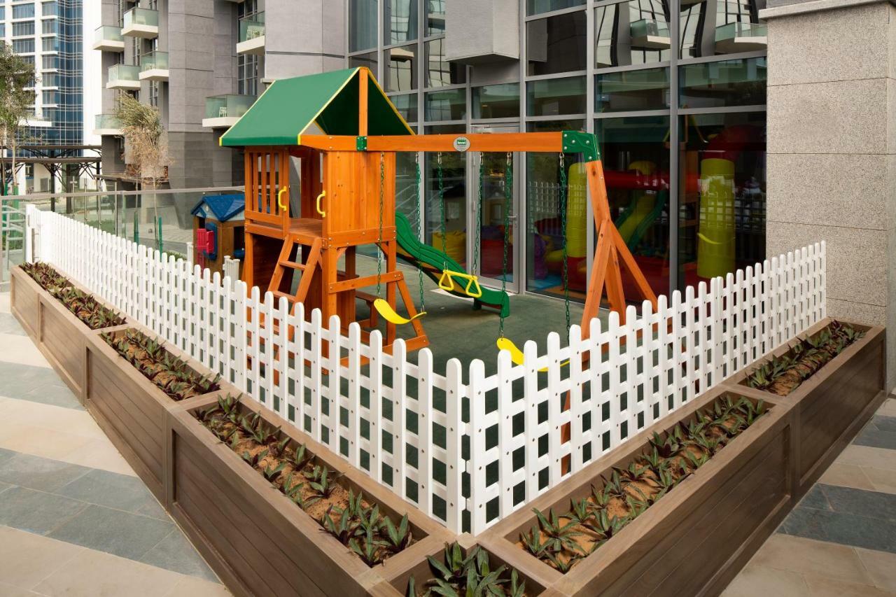두바이 밀레니엄 아트리아 비즈니스 베이 아파트 호텔 외부 사진 The image shows a children's playground area, which is enclosed by a white picket fence. The playground features a wooden play structure with a slide, swings, and climbing elements. Surrounding the play area are planters with greenery, and the backdr