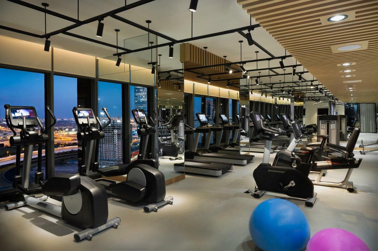 두바이 밀레니엄 아트리아 비즈니스 베이 아파트 호텔 외부 사진 The photo shows a modern gym with various types of exercise equipment available. There are several treadmills and elliptical machines arranged in rows, with large windows in the background providing a view of the city. The lighting is bright and cont