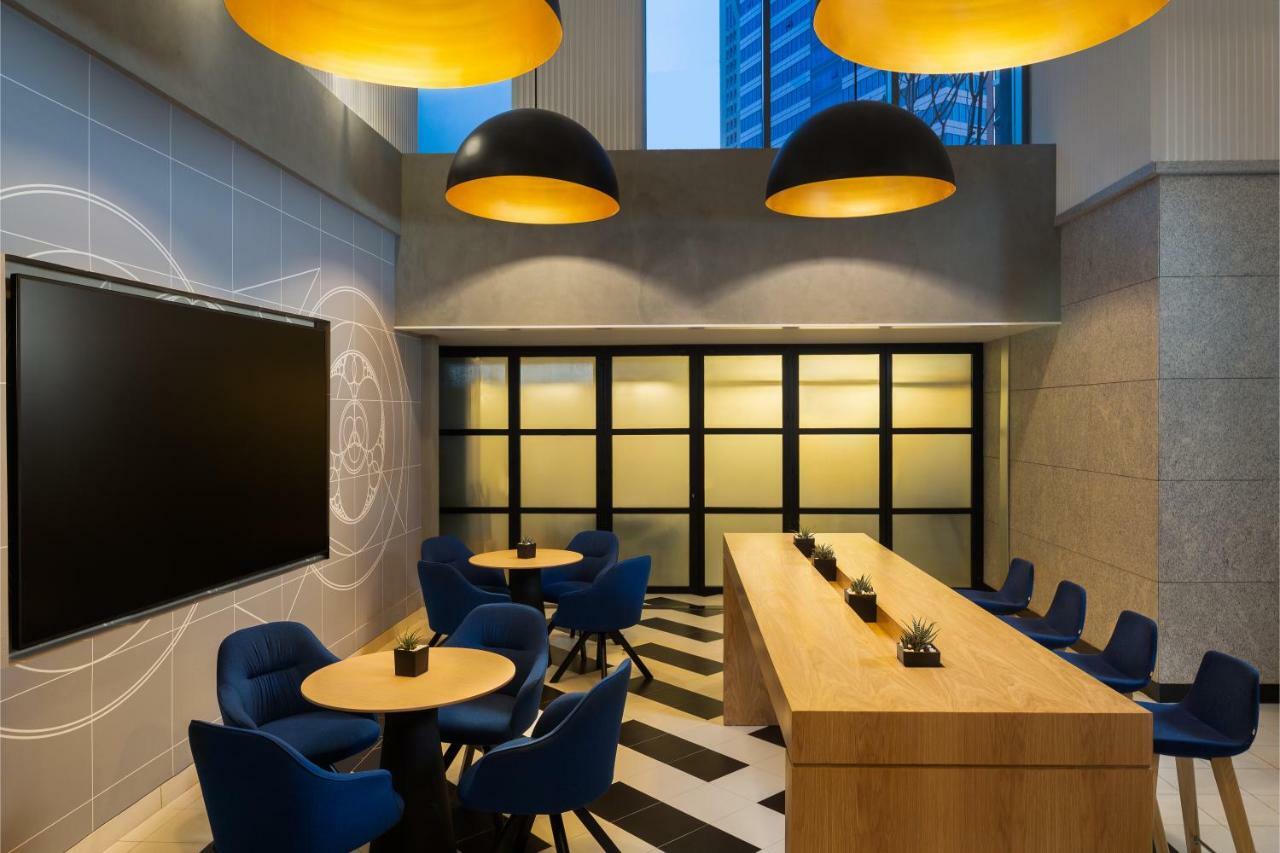 두바이 밀레니엄 아트리아 비즈니스 베이 아파트 호텔 외부 사진 The photo shows a modern interior space, likely a lounge or meeting area. It features a long wooden table with some small plants on it, accompanied by several blue, circular chairs arranged around it. There is a large flat-screen television mounted o