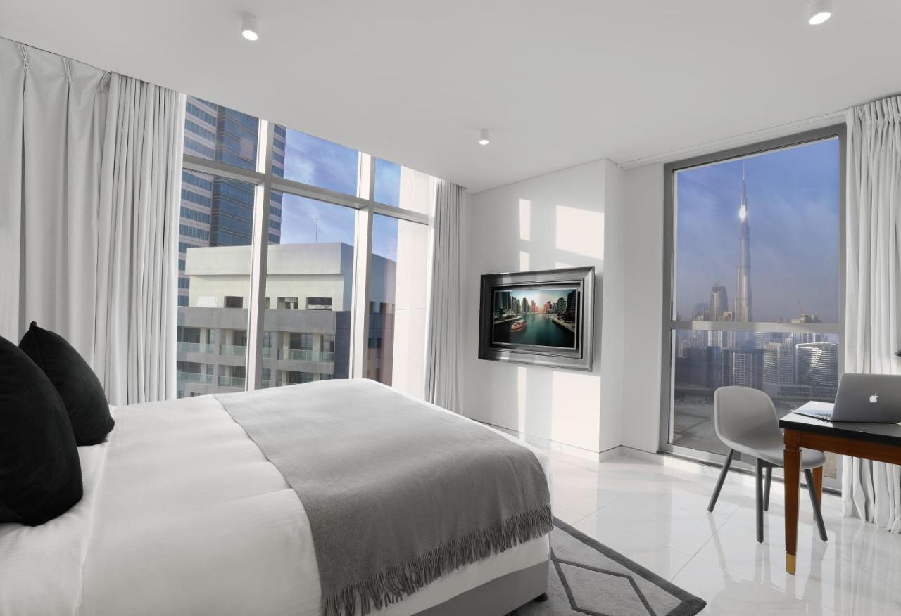 두바이 밀레니엄 아트리아 비즈니스 베이 아파트 호텔 외부 사진 The photo depicts a modern hotel room that features a large bed with a gray throw blanket. There are large windows allowing natural light to fill the space, showcasing a city skyline view. A flat-screen television is mounted on the wall opposite the 